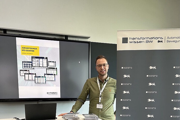 Ein Mann steht zwischen Monitor und Roll-up und blickt lächelnd in die Kamera.