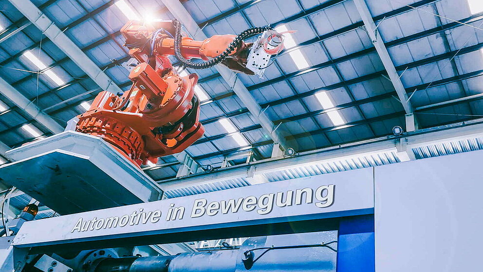 Roboterarm sitzt erhöht in einer Produktionshalle. Auf der Anlage steht "Automotive in Bewegung" geschrieben.