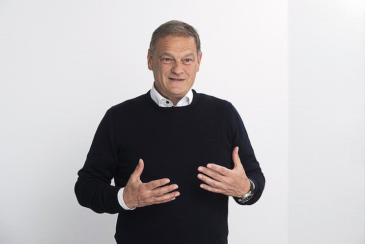 Ein Mann mit schwarzem Pulli blickt lächelnd in die Kamera mit offener Gestik.